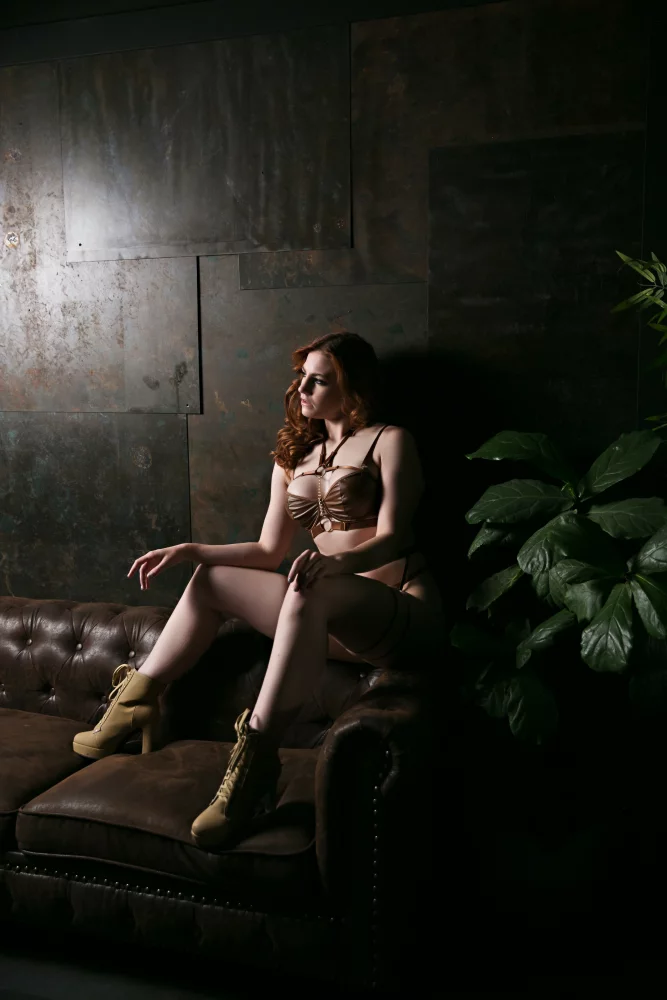 Image of Boudoir portrait of a woman in leather lingerie surrounded by moody lighting and lush greenery.